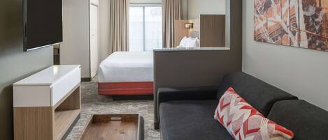 A hotel room with a flat-screen TV, a bed, a sofa, a wooden side table, and a large abstract painting on the wall.