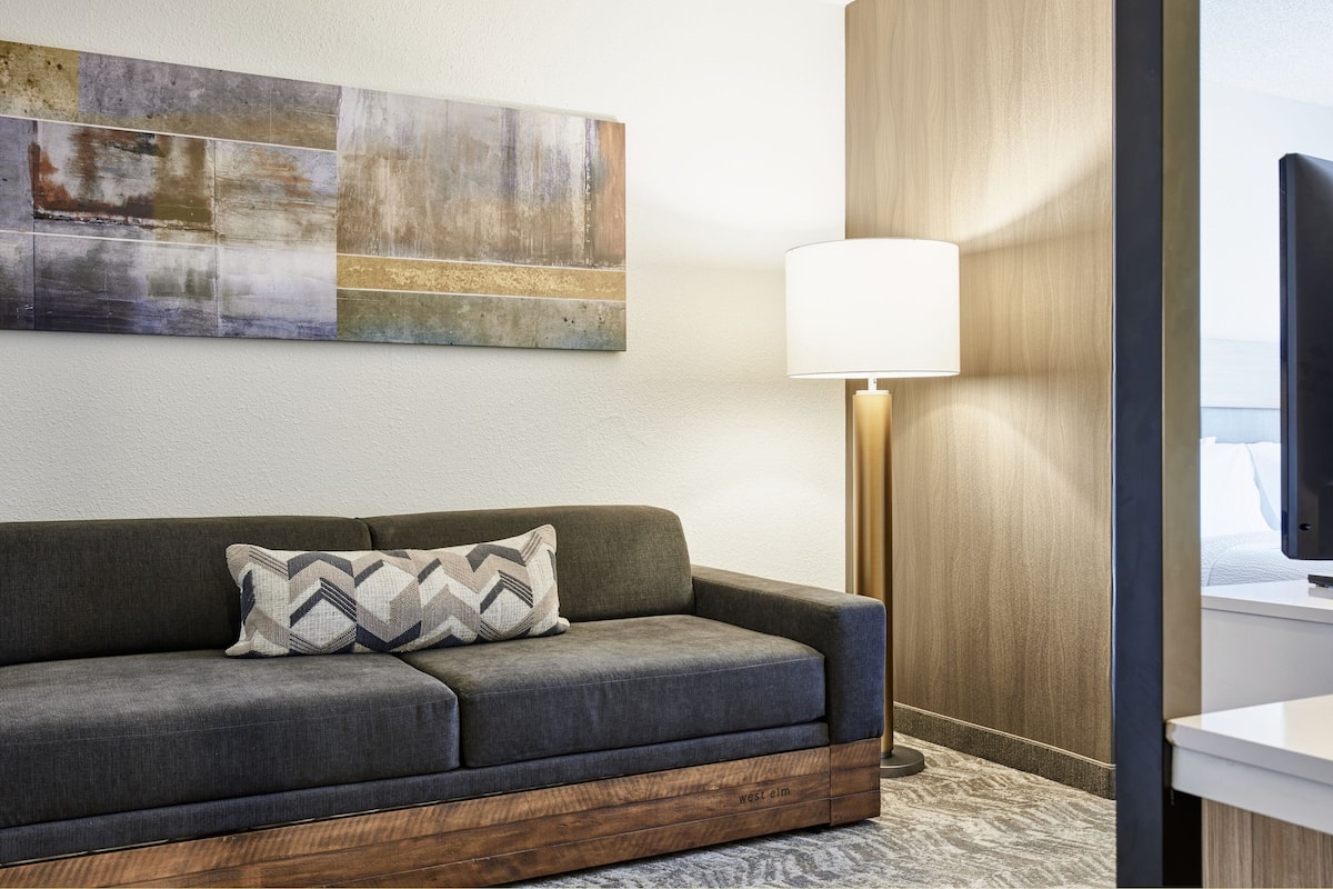 A modern hotel room with a gray sofa, a wooden side table, a large abstract painting, a floor lamp, and a television.