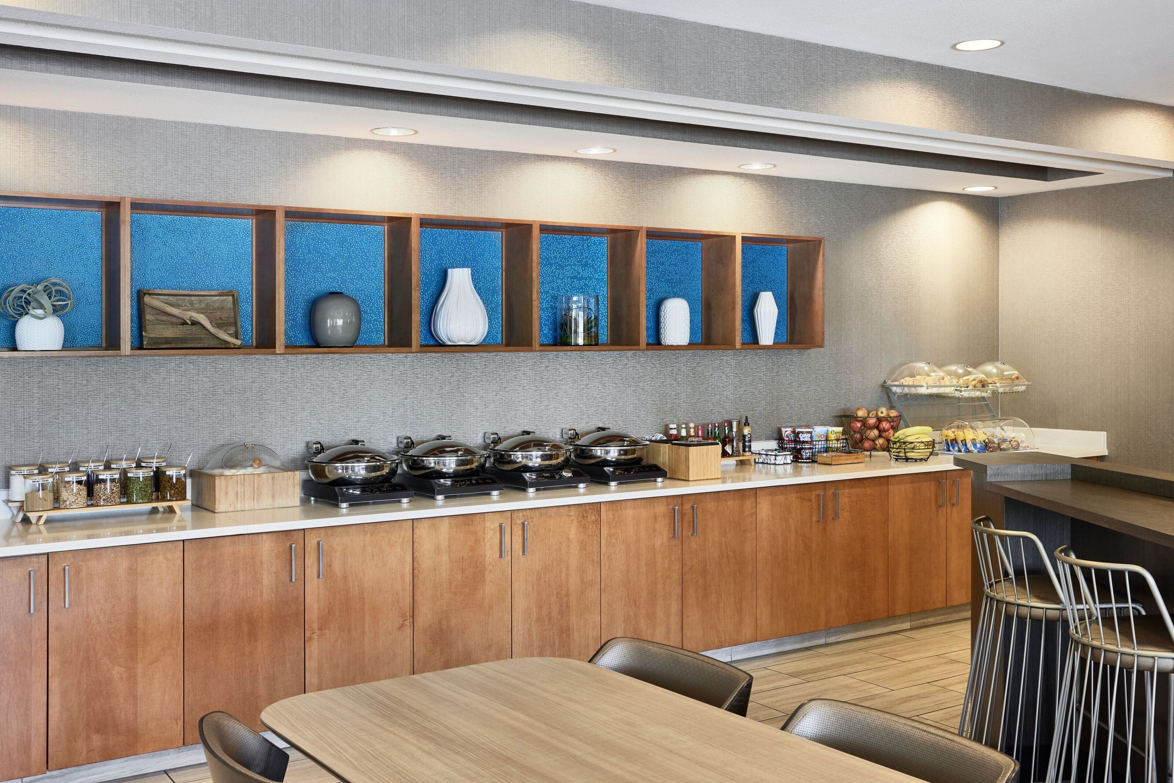 A modern kitchen with wooden cabinets, a central island with cooking appliances, and a breakfast bar with food items.