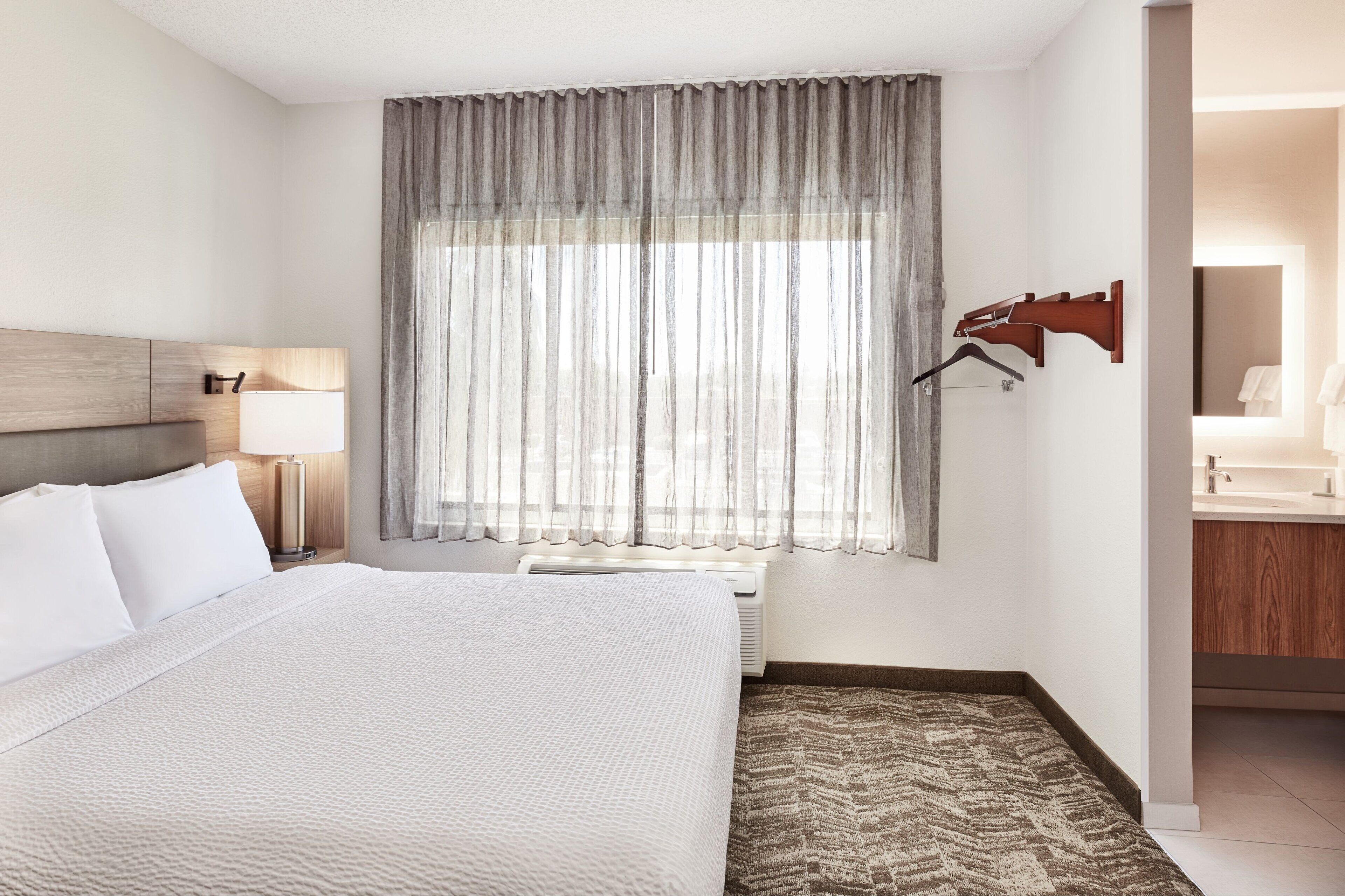 A hotel room with a large bed, a bedside lamp, a window with sheer curtains, a wall-mounted coat rack, and a bathroom visible through an open door.