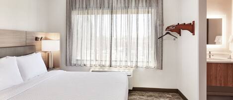 A hotel room with a large bed, a bedside lamp, a window with sheer curtains, a wall-mounted coat rack, and a bathroom visible through an open door.