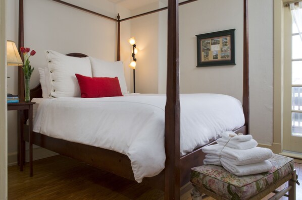 A four-poster bed with white linens and a red accent pillow, a bedside table with a lamp and a vase of flowers, and a window with a view of buildings.