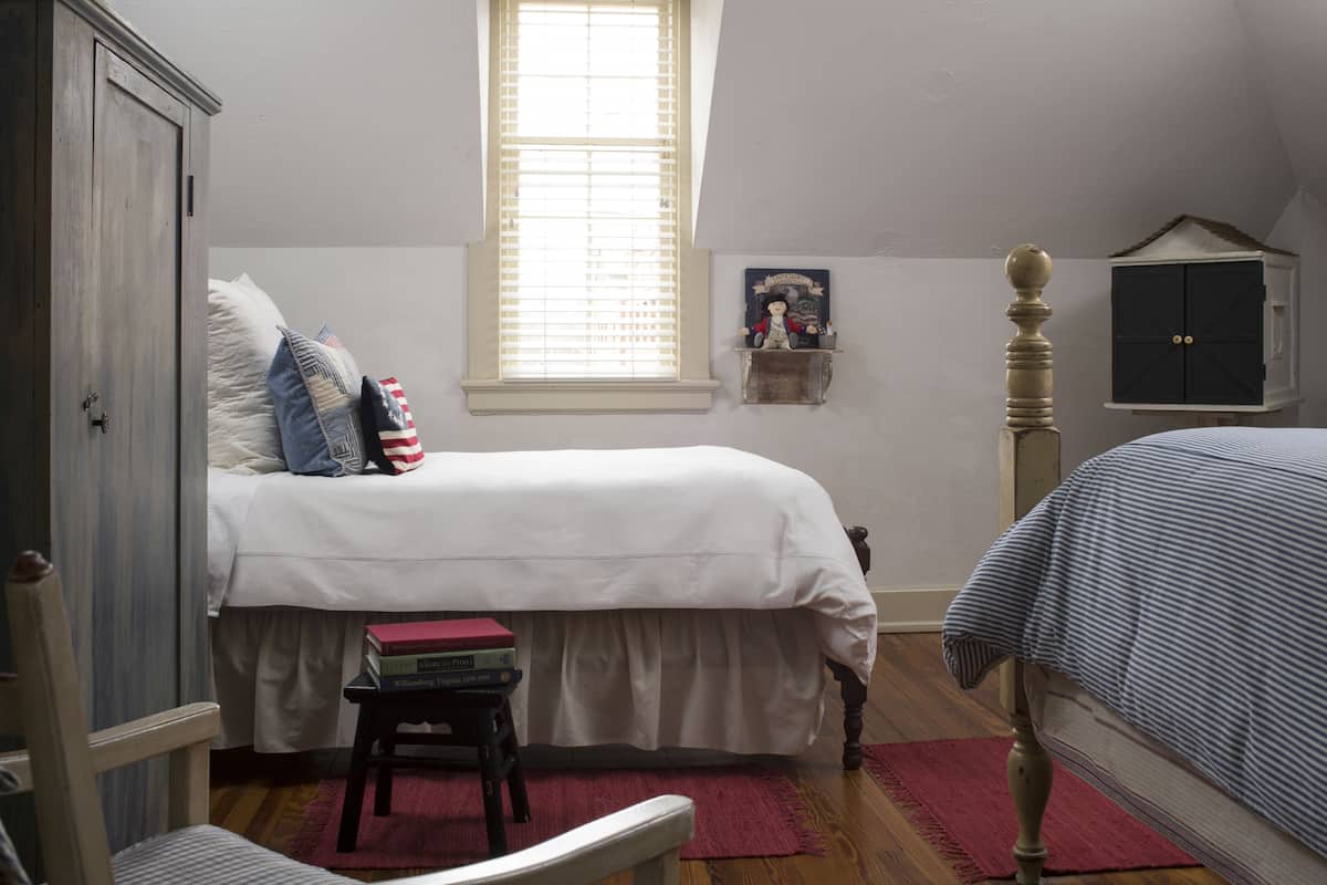 A bedroom with a bed, a small table with books, a window, and a wardrobe.