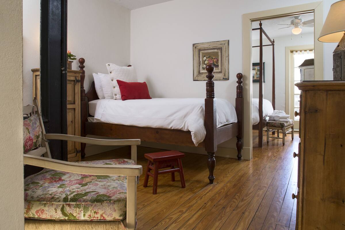 A bedroom with a four-poster bed, a wooden chair, a small stool, and a wooden cabinet.