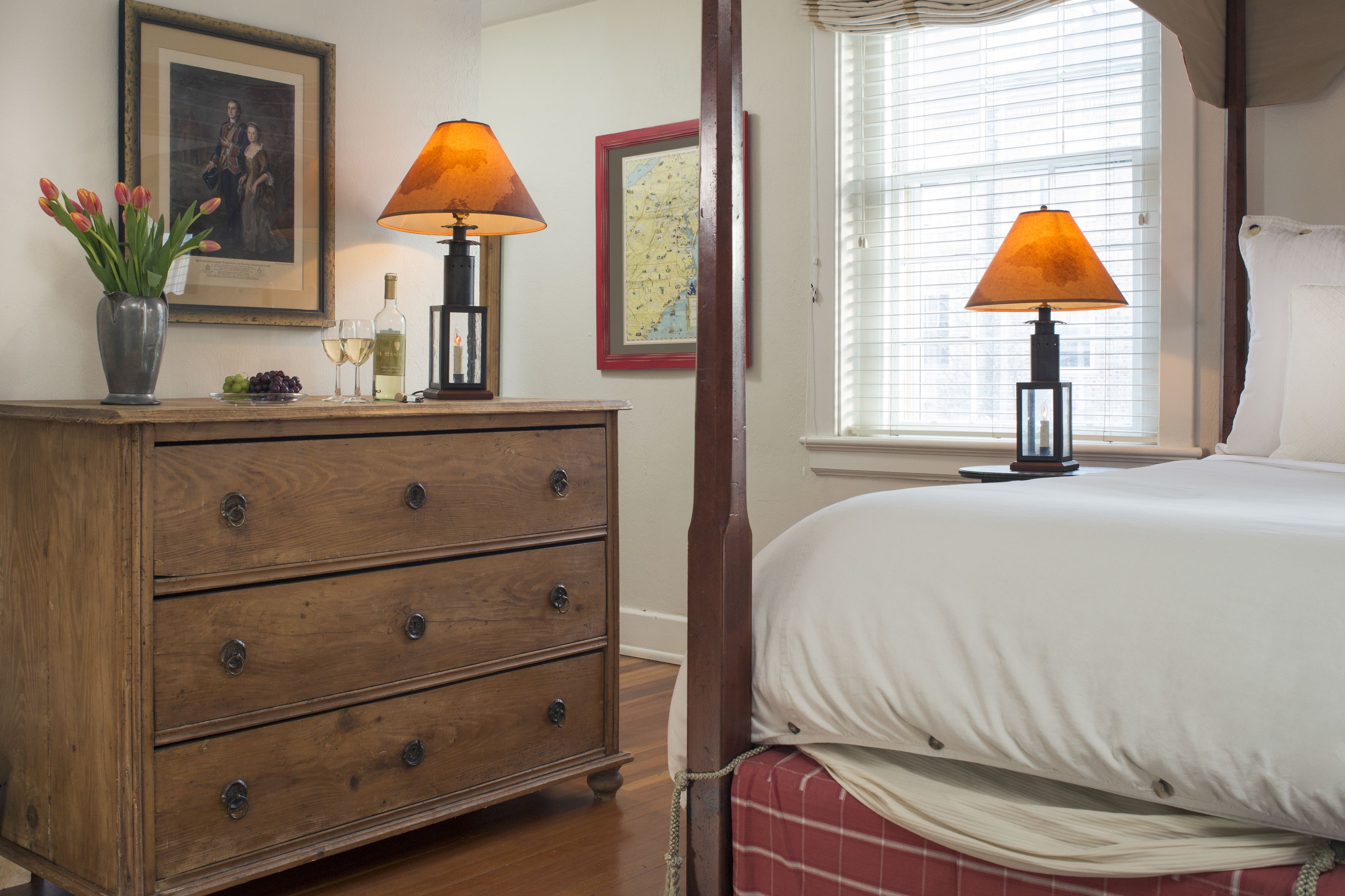 A bedroom with a wooden dresser, two lamps, a bed with white bedding, a window with blinds, and a framed map on the wall.