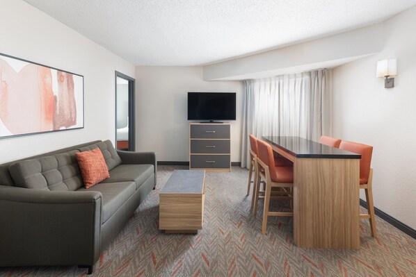 Una habitación de hotel moderna con un sofá gris, una mesa de centro de madera, un televisor, una mesa de comedor con sillas color naranja y una alfombra estampada.