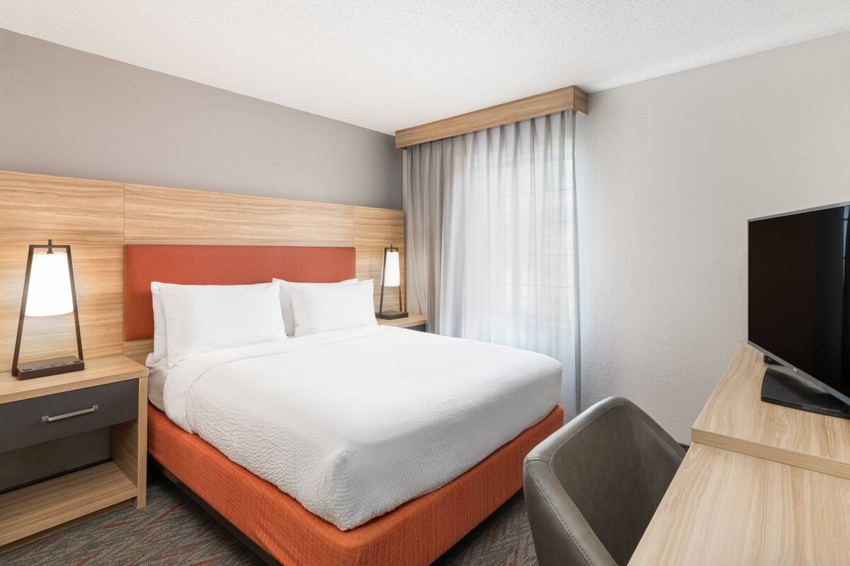 A hotel room with a bed, a desk with a TV, and a nightstand with a lamp.