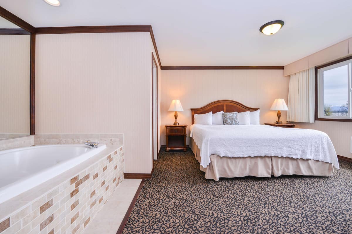 Habitación de hotel con una bañera grande, una cama doble con ropa blanca, dos lámparas de noche y una alfombra estampada.