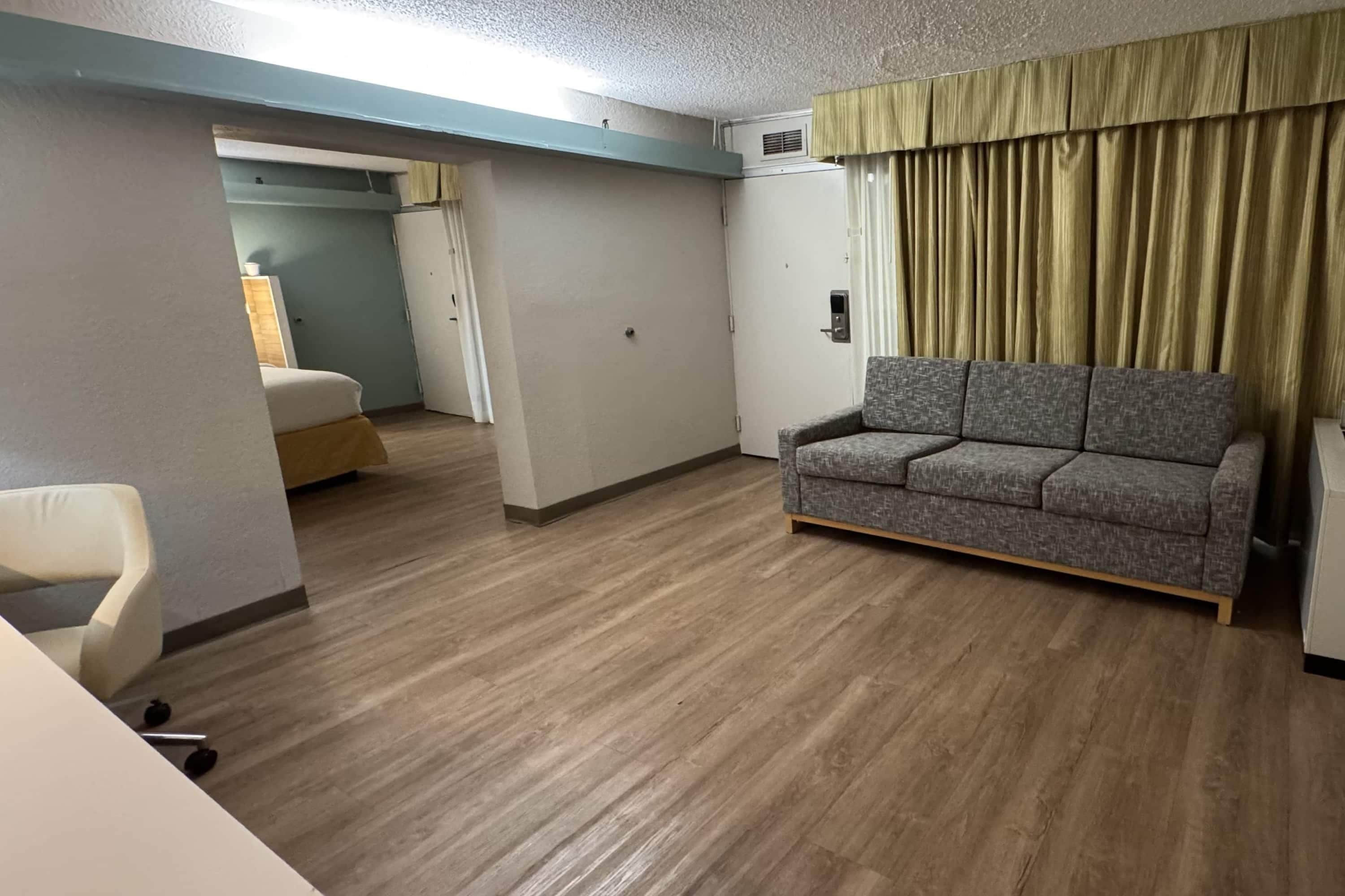 Habitación de hotel con sofá gris, una silla blanca, cama y suelo de madera.