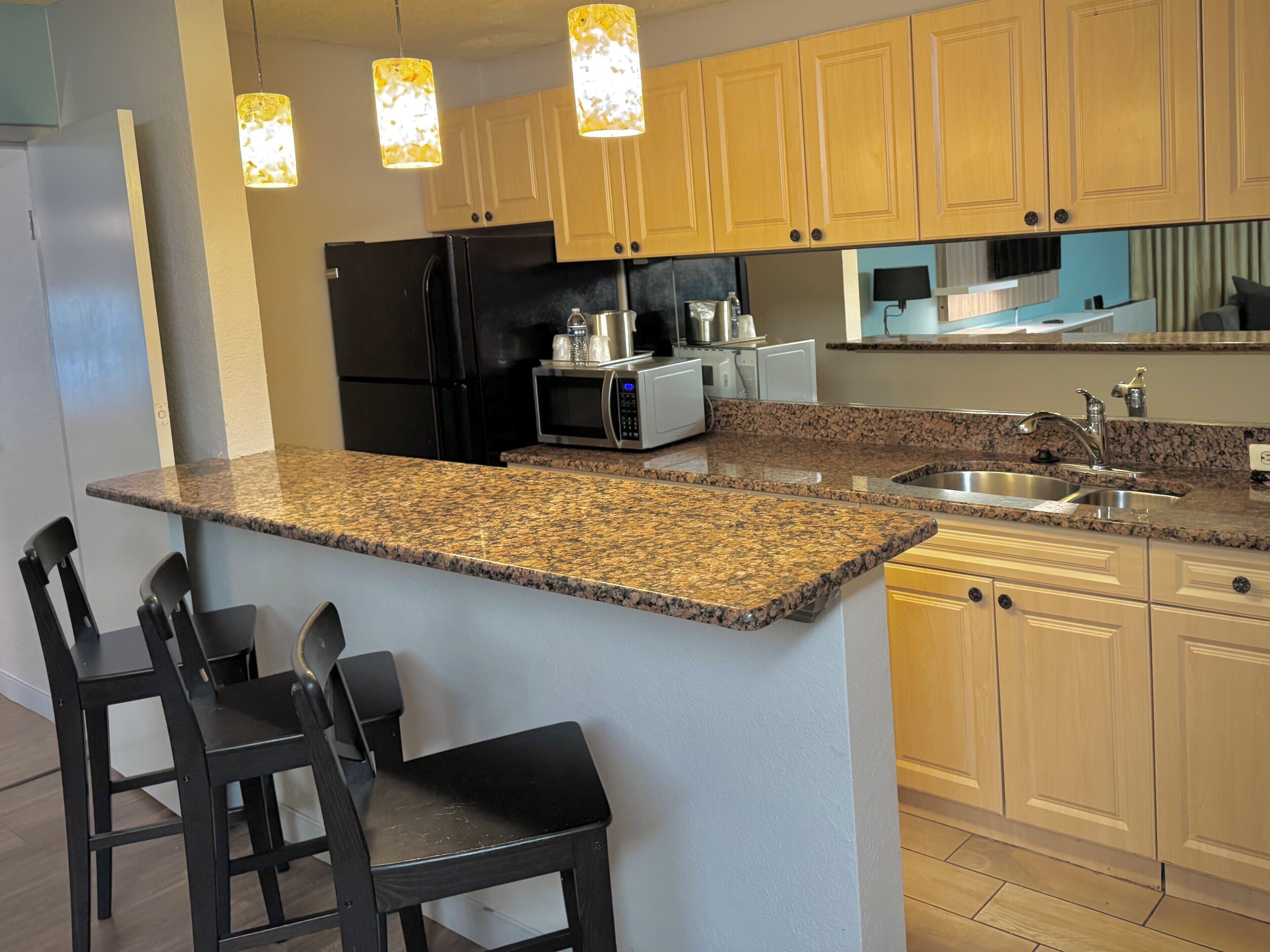 A kitchen with granite countertops, wooden cabinets, a black refrigerator, a microwave, a sink, and three black bar stools.