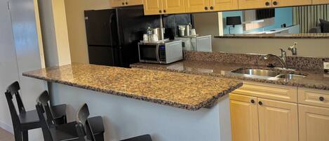 A kitchen with granite countertops, wooden cabinets, a black refrigerator, a microwave, a sink, and three black bar stools.