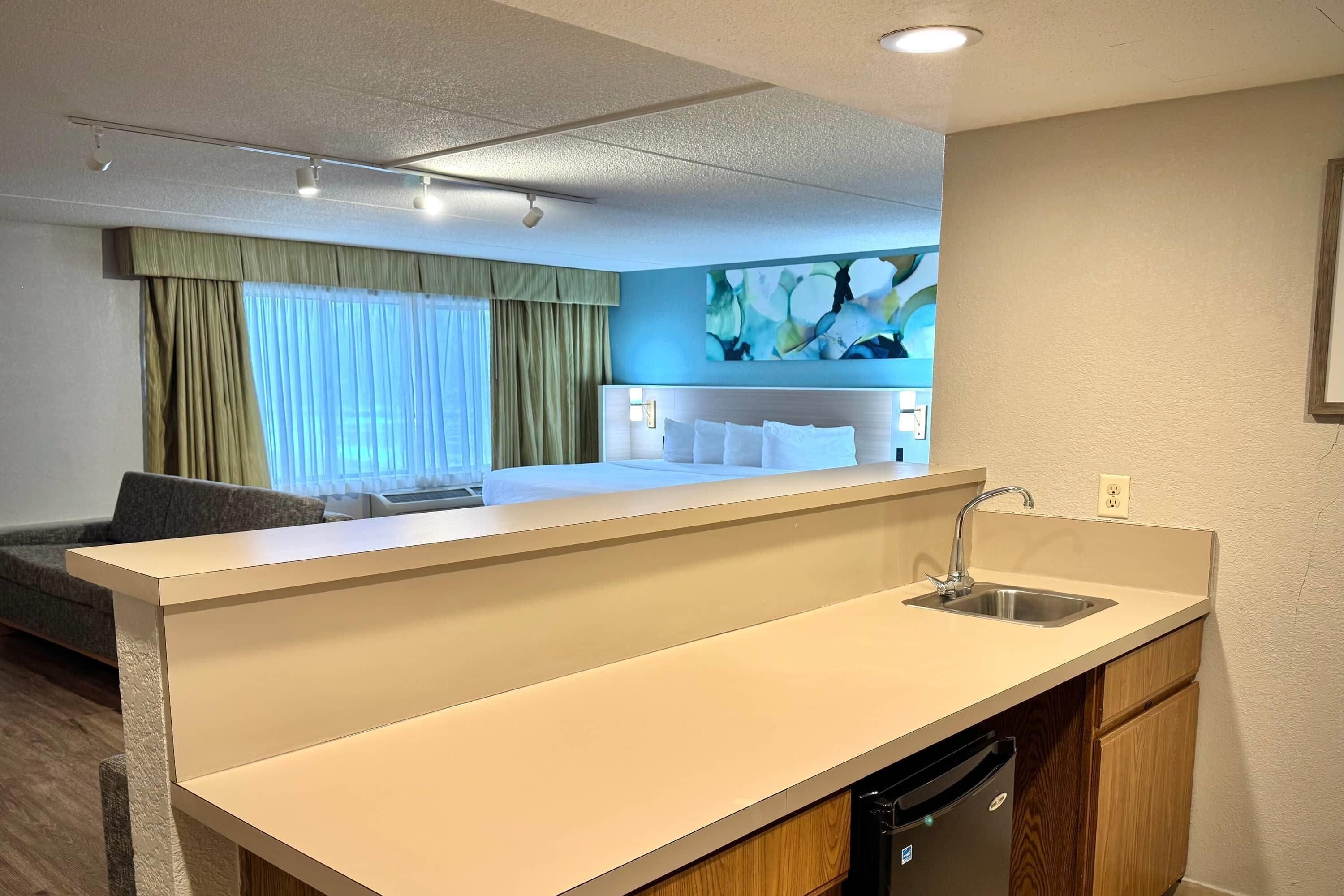 A hotel room with a kitchenette, a bed with white linens, a gray sofa, and a large abstract painting on the wall.