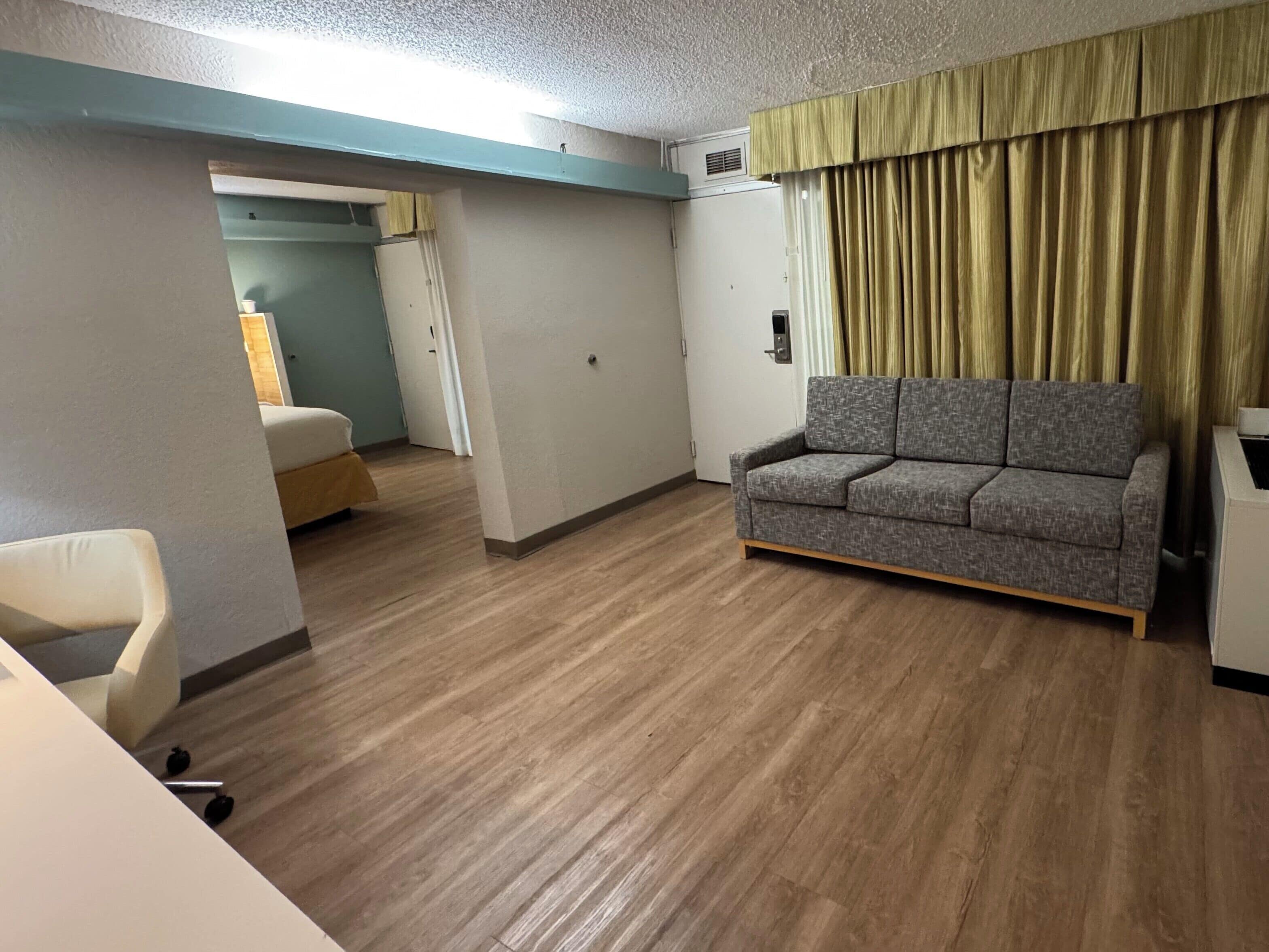 Una habitación con un sofá gris, una silla blanca y suelo de madera. Hay una puerta que da a otra habitación.