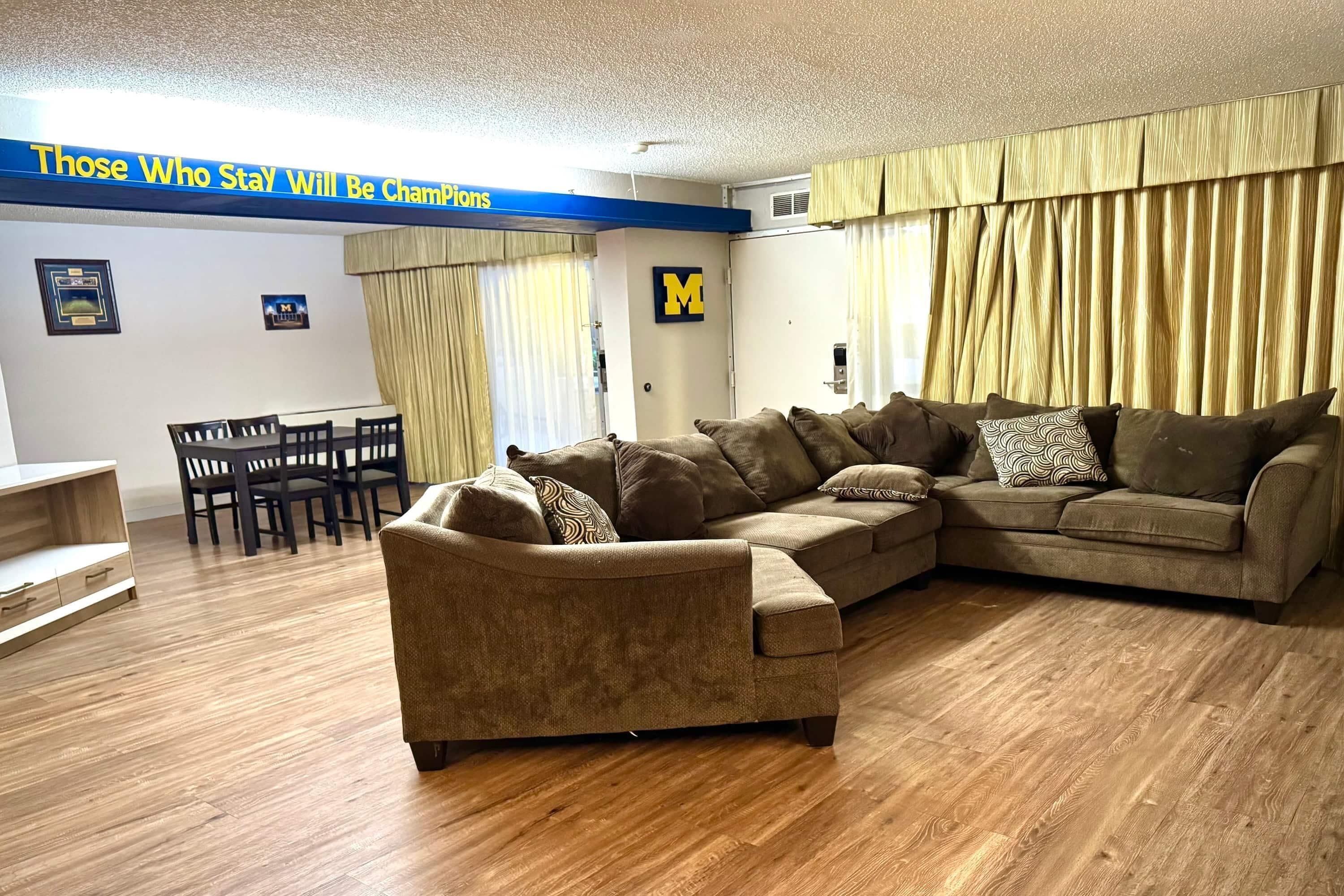 Uma sala de estar com sofá em formato de L, uma mesa de jantar com cadeiras e uma janela grande com cortinas.