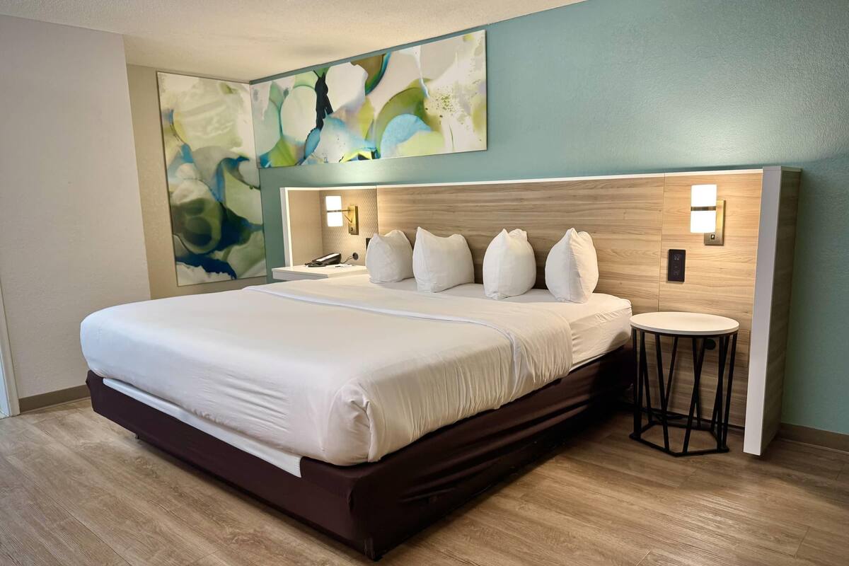 Ein modernes Hotelzimmer mit einem großen Bett, einem Kopfteil mit integrierter Beleuchtung, einem kleinen runden Tisch und abstrakter Wandkunst.