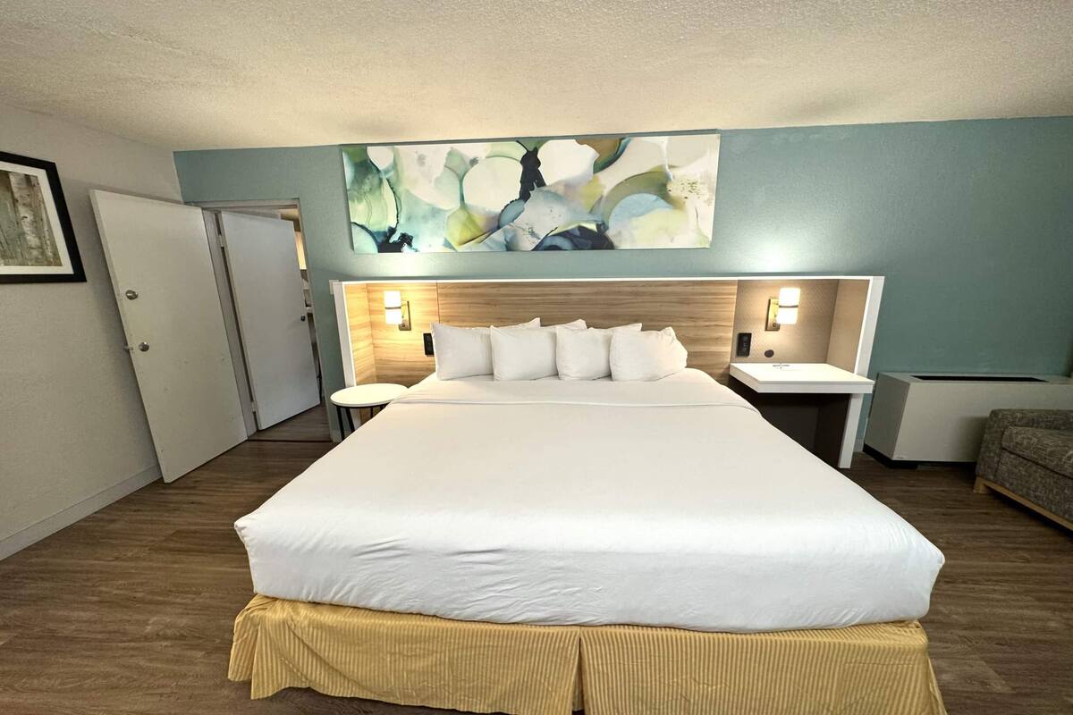 Ein Hotelzimmer mit einem großen Bett, zwei Nachttischen, einem Bettrahmen mit integrierter Beleuchtung, einem Gemälde über dem Bett und einer Tür links.