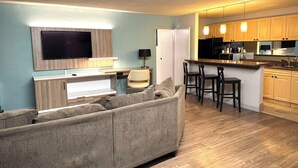 Un salon moderne comprenant un canapé, un téléviseur fixé au mur, un bureau et un coin cuisine avec des tabourets de bar.