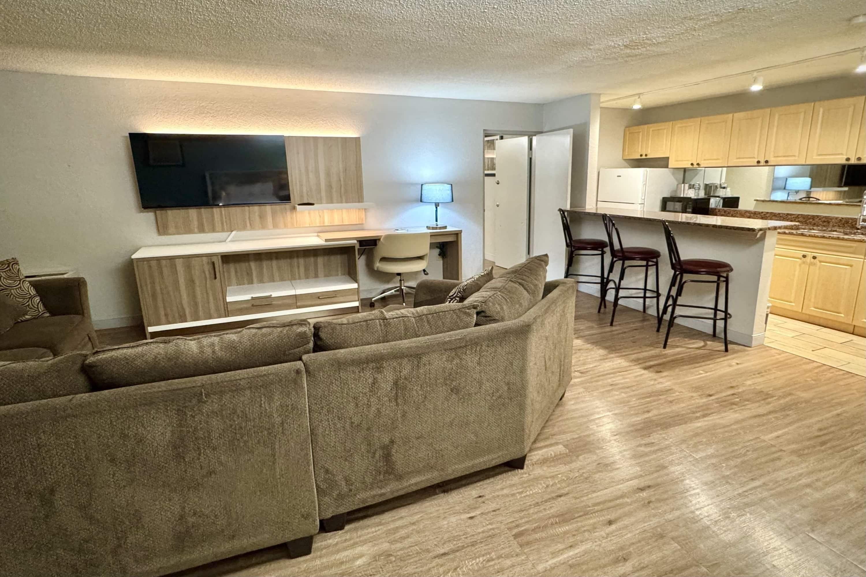 A modern living room with a sofa, a TV mounted on the wall, a desk with a chair, and a kitchen area with wooden cabinets and a bar stool.