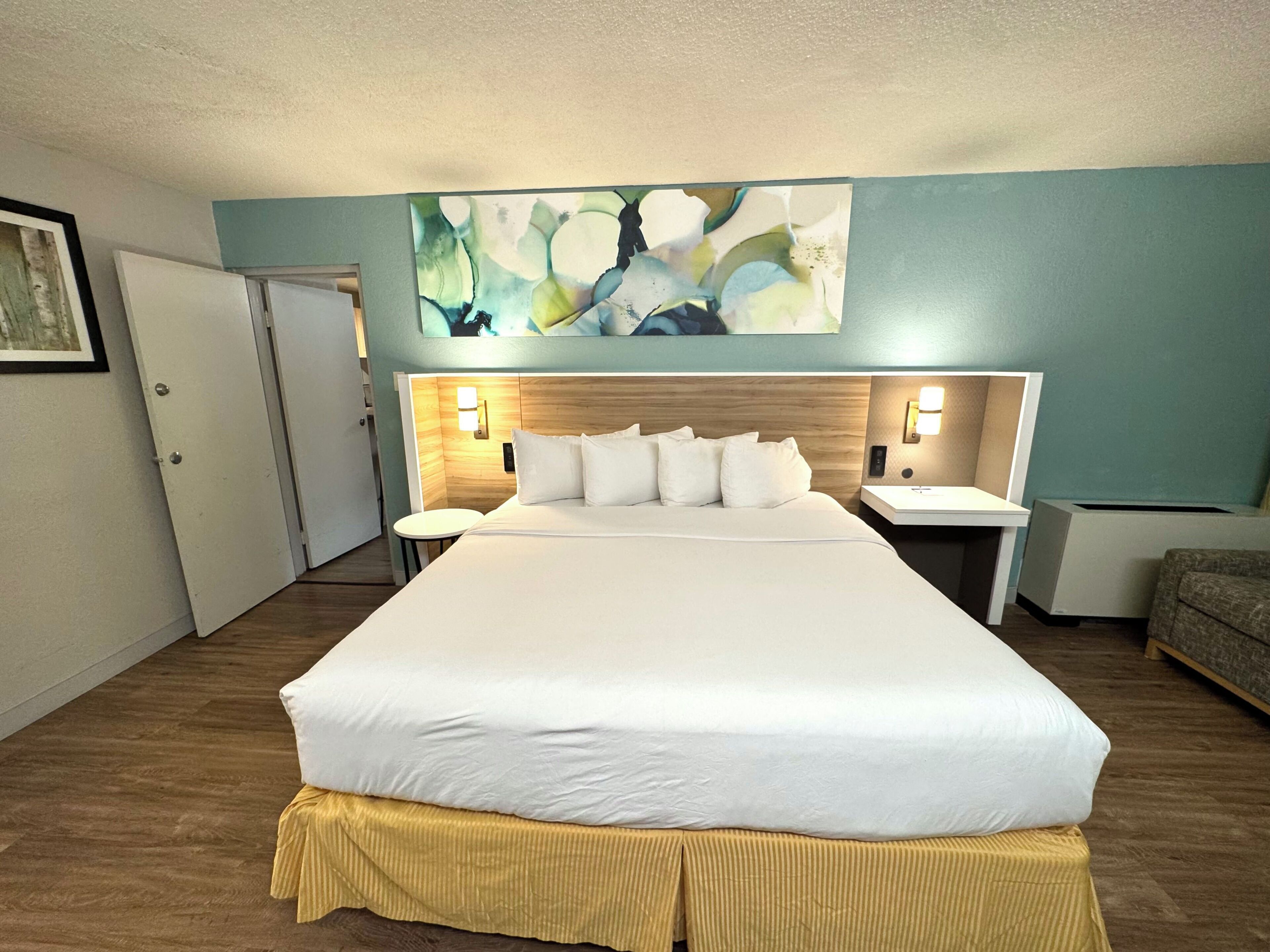 A hotel room with a large bed, two bedside tables, a headboard, a wall-mounted artwork, and a door.