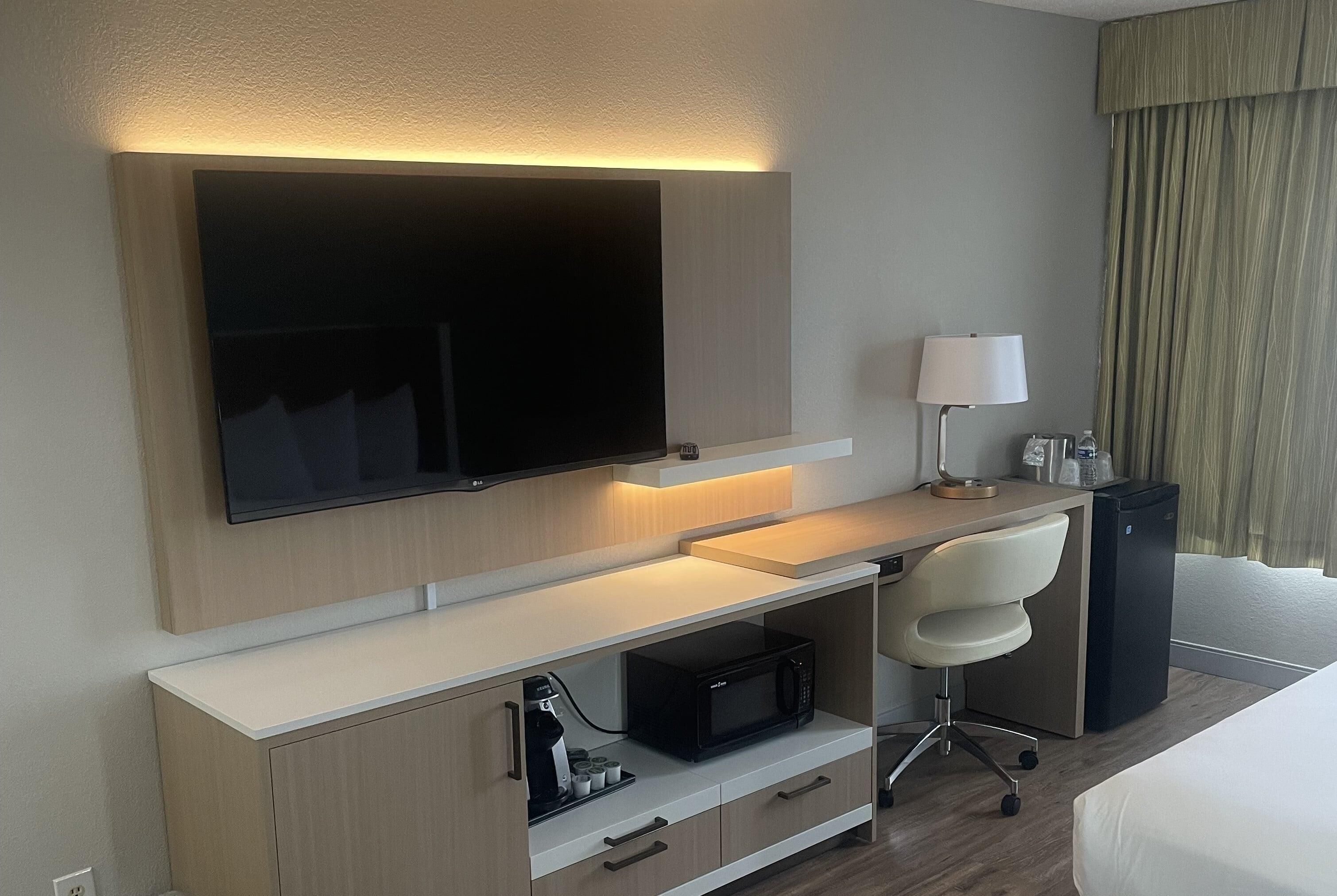 A hotel room with a flat-screen TV mounted on the wall, a desk with a lamp, a microwave, and a small refrigerator.