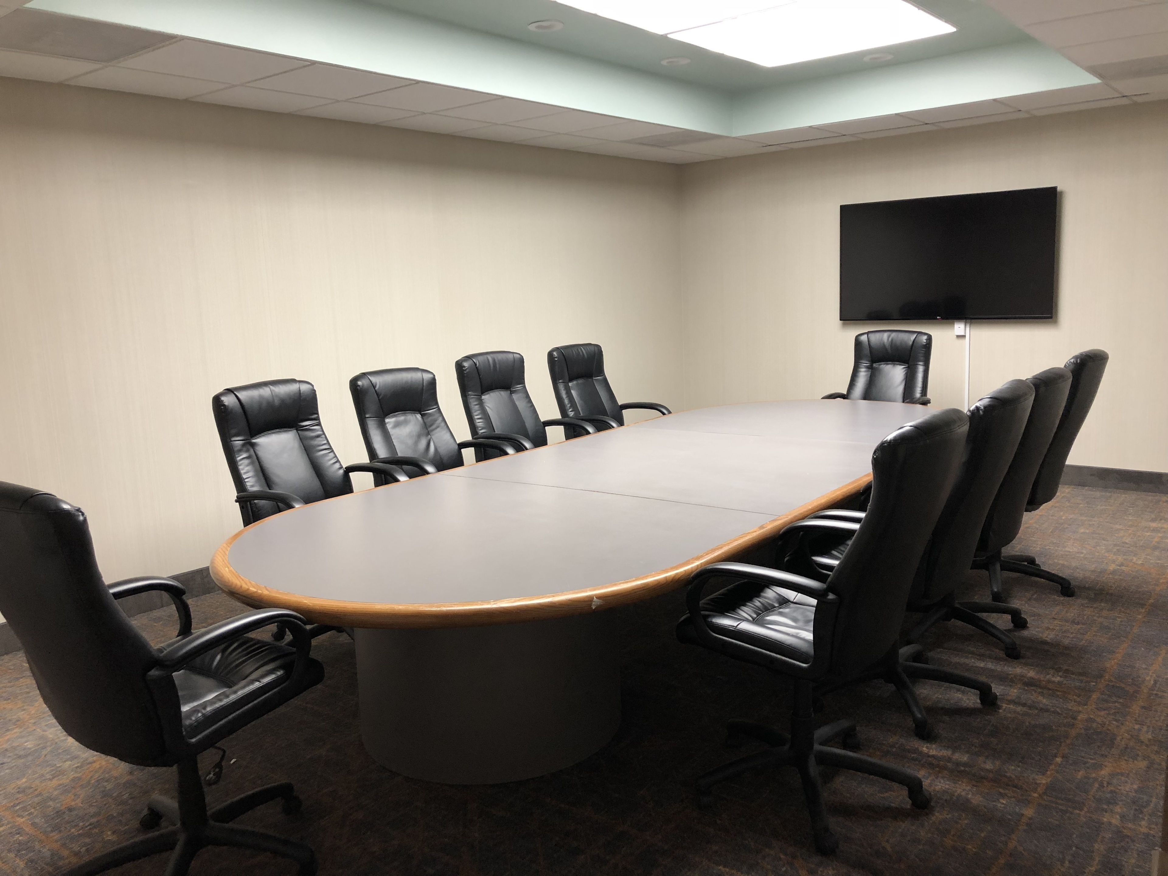 A conference room with a long table, black leather chairs, a flat-screen TV, and a ceiling light.