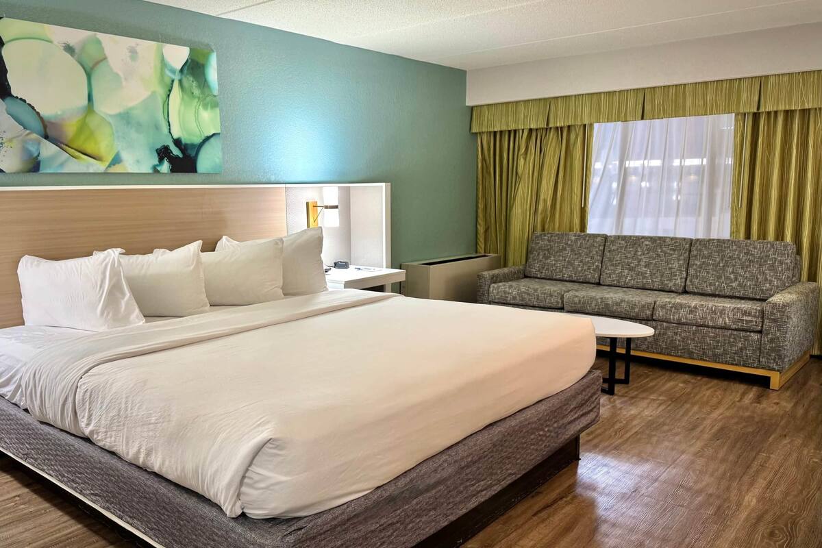 Ein Hotelzimmer mit einem großen Bett, einem grauen Sofa, einem kleinen Tisch und einem Gemälde an der Wand.