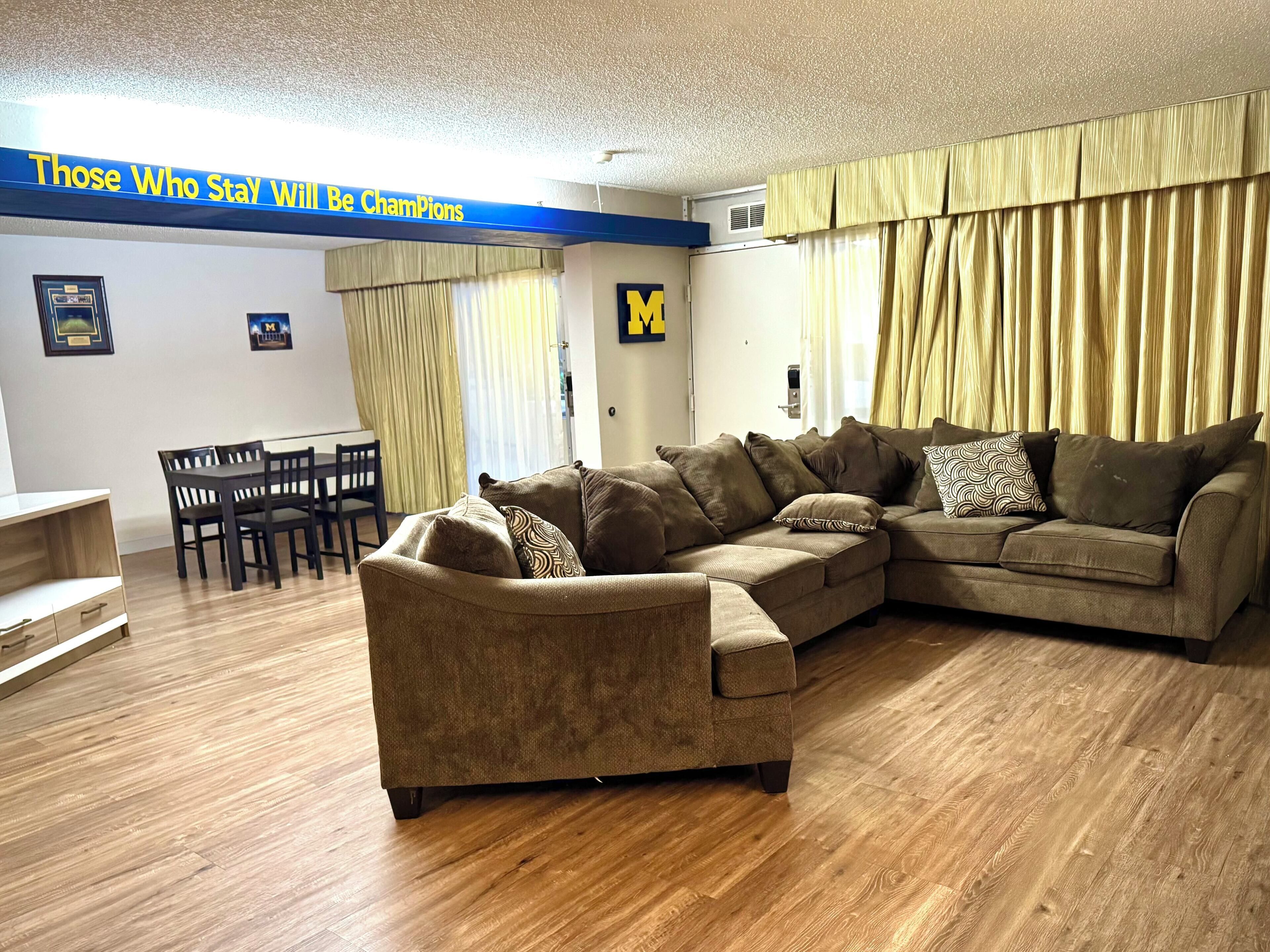 A living room with a sectional sofa, wooden flooring, a dining table with chairs, and a wall with a motivational quote.