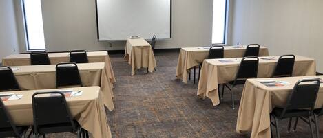 Un salón de conferencias con mesas, sillas, una pantalla de proyector y ventanas.
