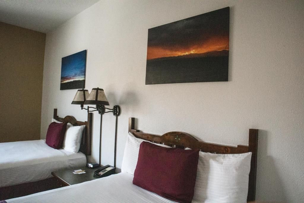 A hotel room with two beds, a nightstand, a lamp, and two paintings on the wall.
