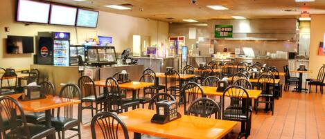 A restaurant with multiple tables and chairs, a counter with a Pepsi refrigerator, and a sign indicating a pick-up order area.