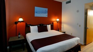 Une chambre d’hôtel avec un grand lit, deux tables de chevet avec des lampes, une tête de lit en bois, un mur rouge et un tableau représentant un paysage enneigé.