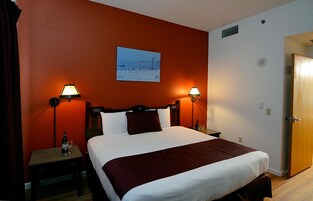 Quarto de hotel com uma cama grande, duas mesas de cabeceira com abajures, cabeceira de madeira, parede vermelha e um quadro de uma paisagem nevada.