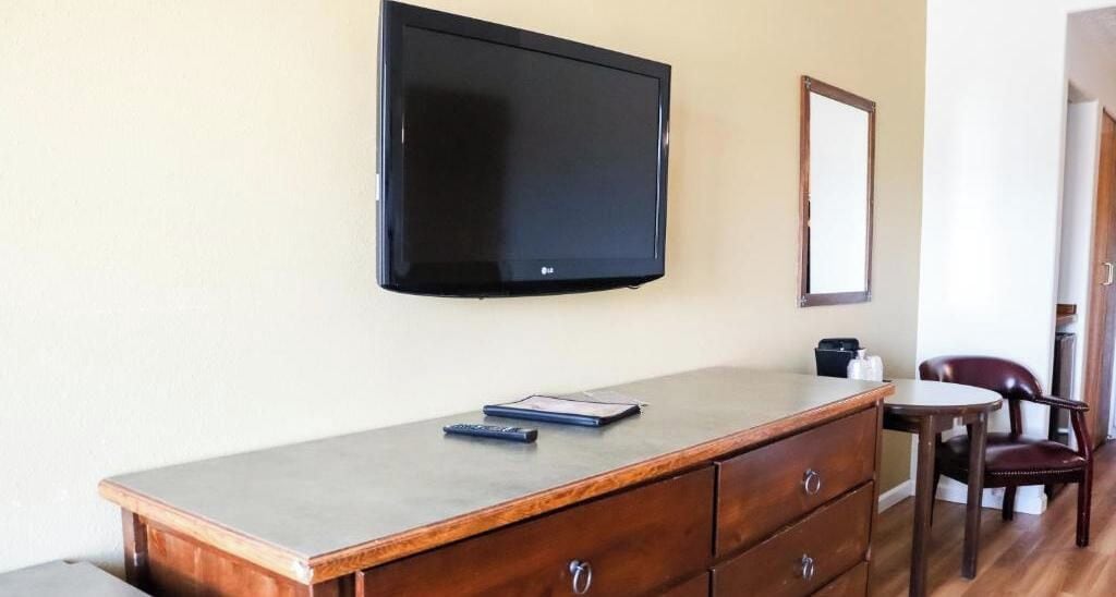 A room with a flat-screen TV mounted on the wall, a wooden desk with drawers, a chair, and a small table.