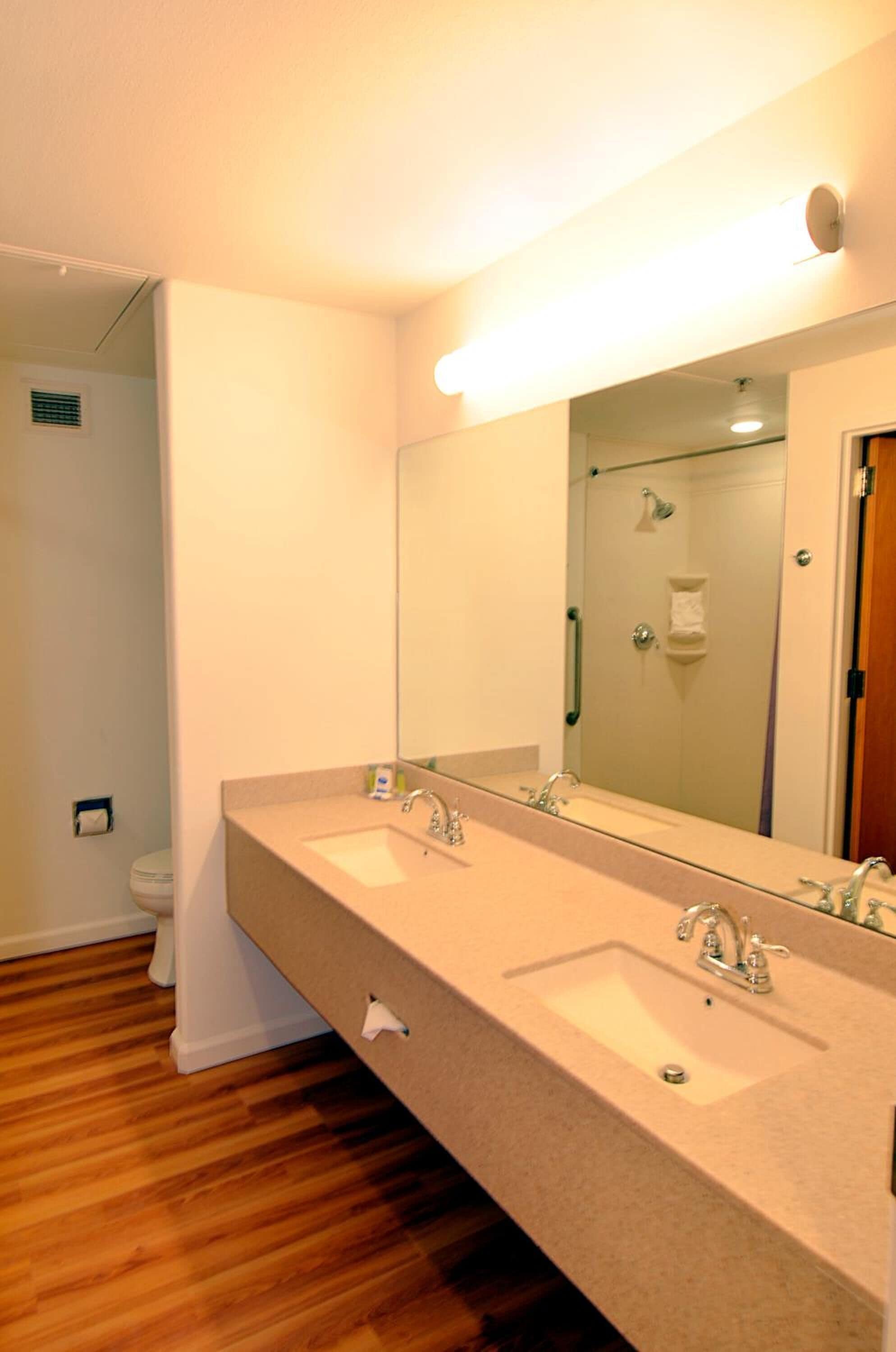 Un baño con lavamanos doble, un espejo grande y una ducha visible a través de un vidrio.