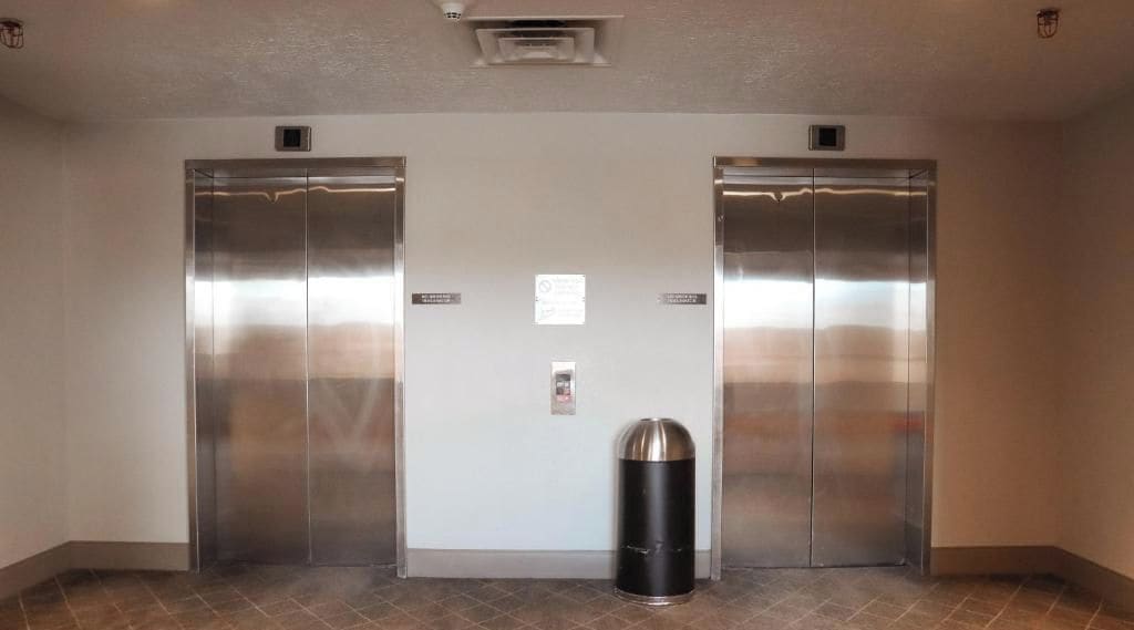 A pair of elevators in a hallway with a trash can in the center.