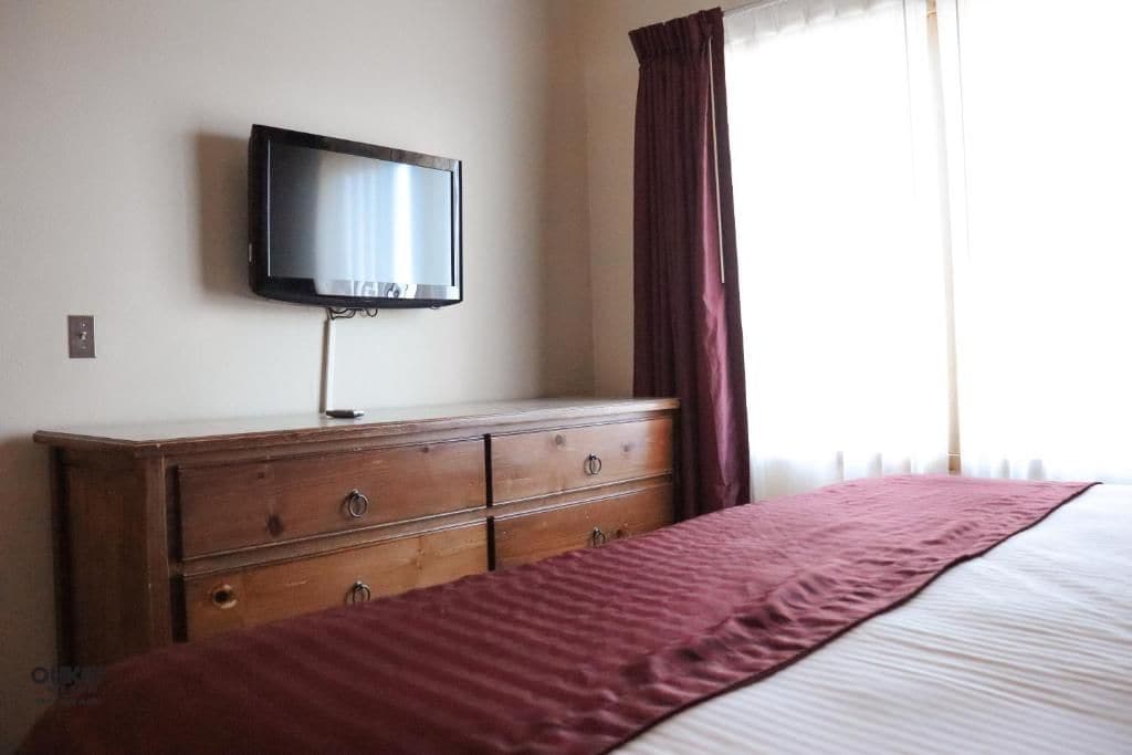 A bedroom with a bed, a wooden dresser, a television mounted on the wall, and a window with curtains.
