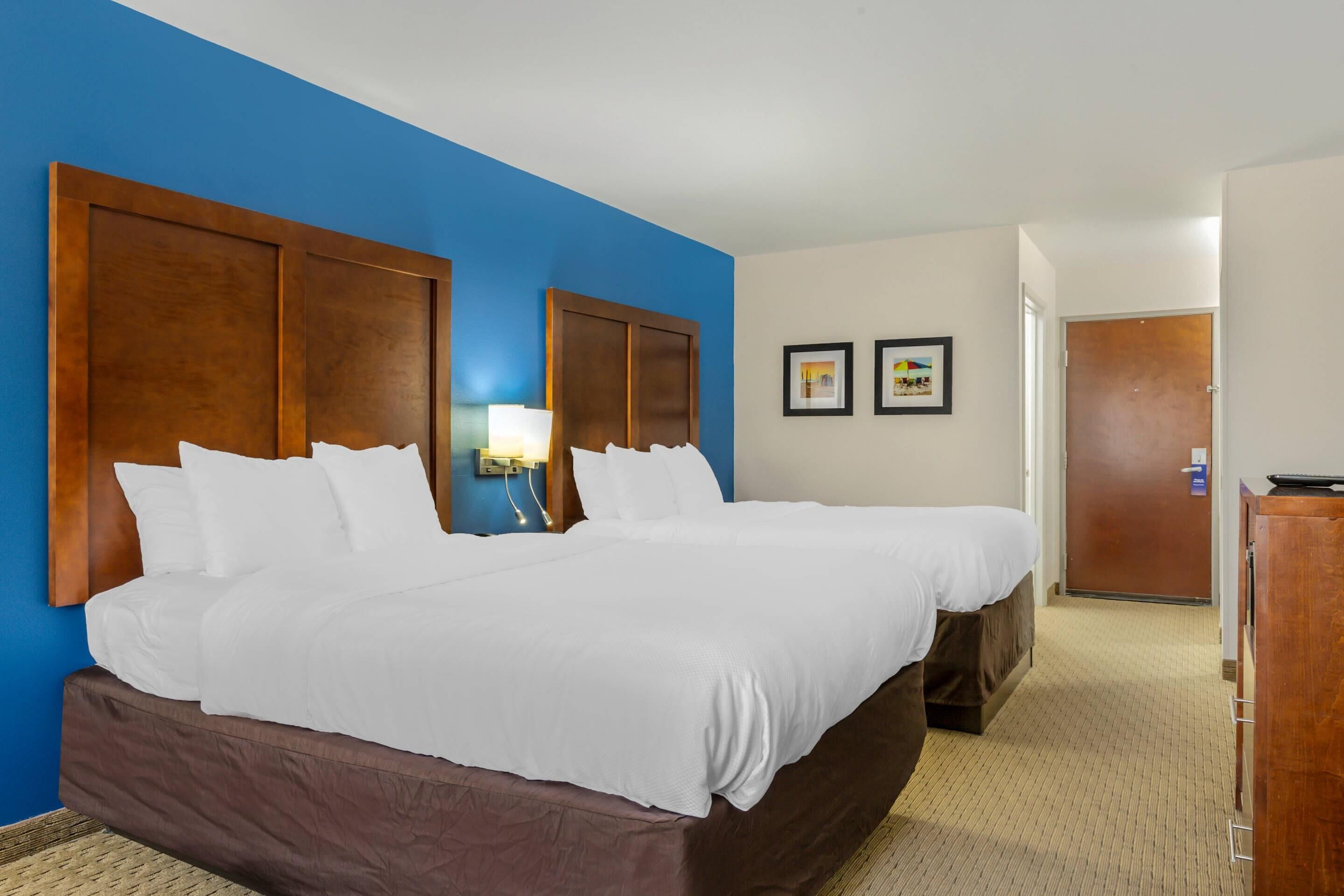 A hotel room with two beds, a wooden headboard, a nightstand, a lamp, and a door.