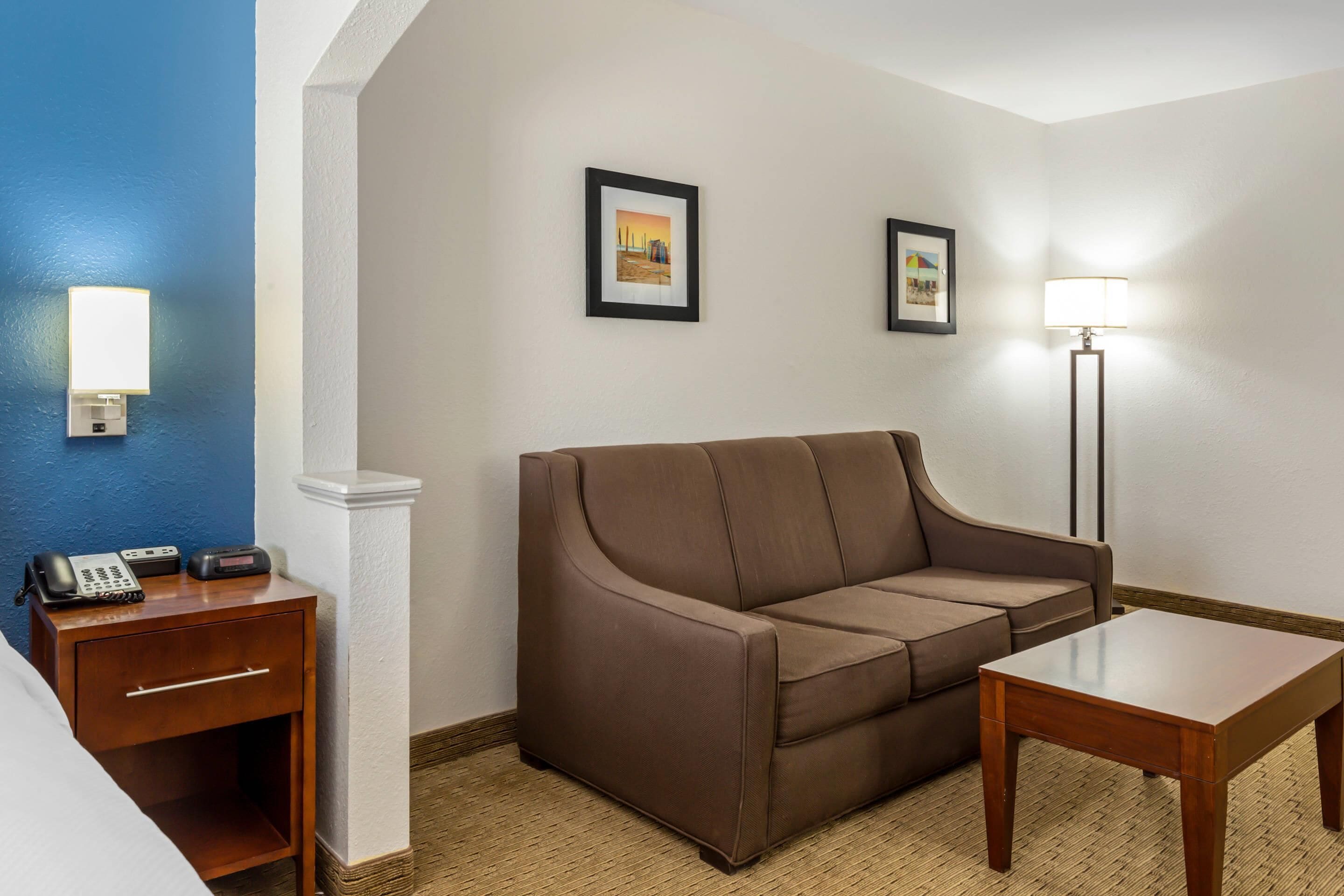 A hotel room with a brown sofa, a wooden nightstand with a phone, a lamp, and two framed pictures on the wall.