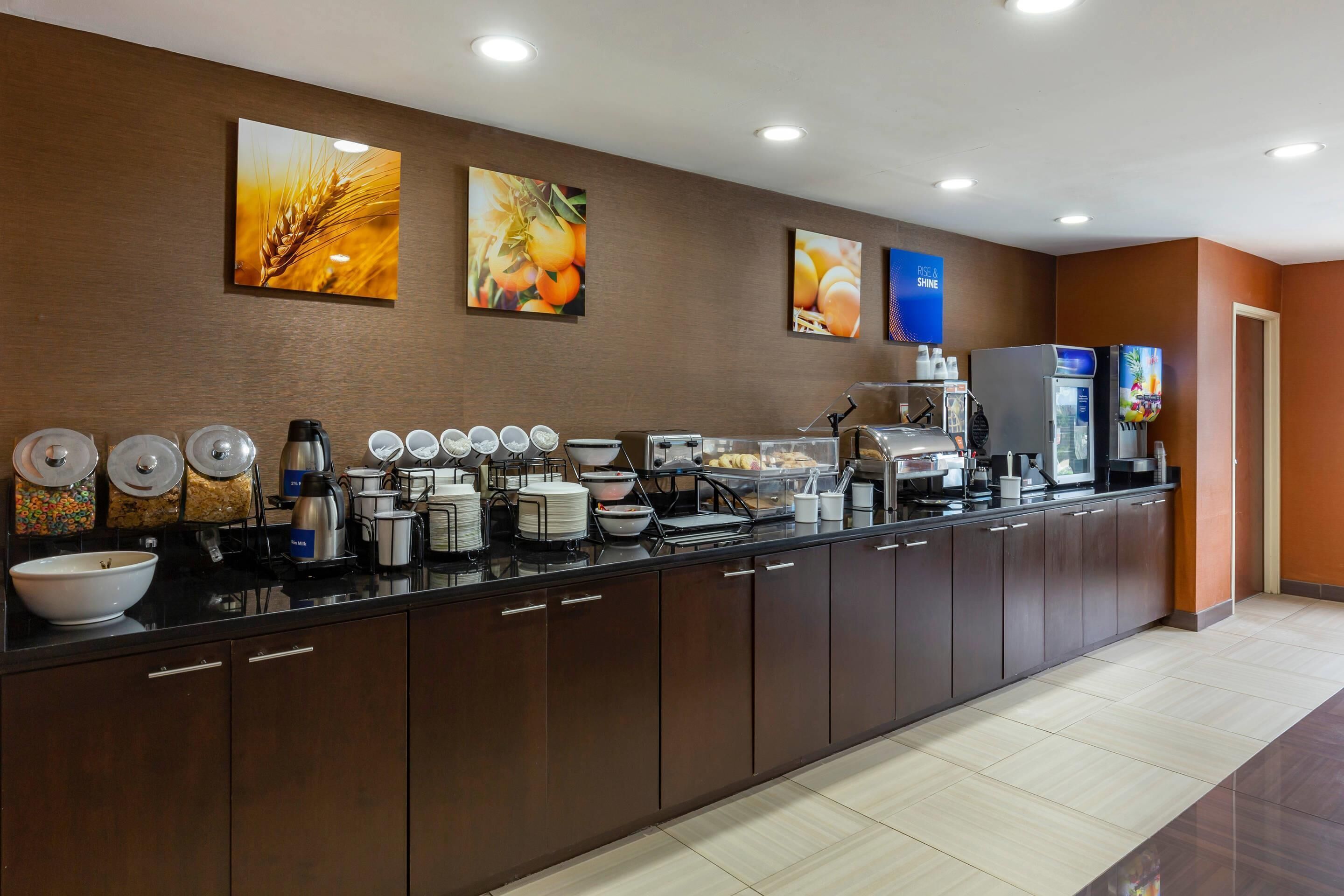 A hotel breakfast area with a long counter, various food items, and artwork on the wall.