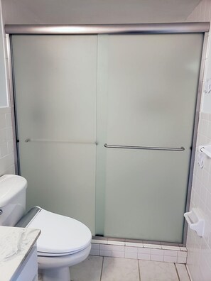 A bathroom with a toilet, a shower enclosure with frosted glass doors, and a white tiled floor. - Comfort Inn Garner Clayton I-40 (Garner)