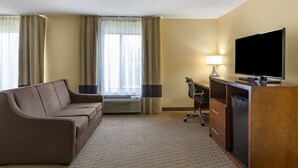 A hotel room with a brown sofa, a desk with a television, a lamp, and a minibar.