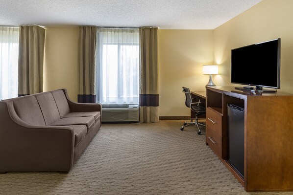 A hotel room with a brown sofa, a desk with a television, a lamp, and a minibar. - Comfort Inn Garner Clayton I-40 (Garner)