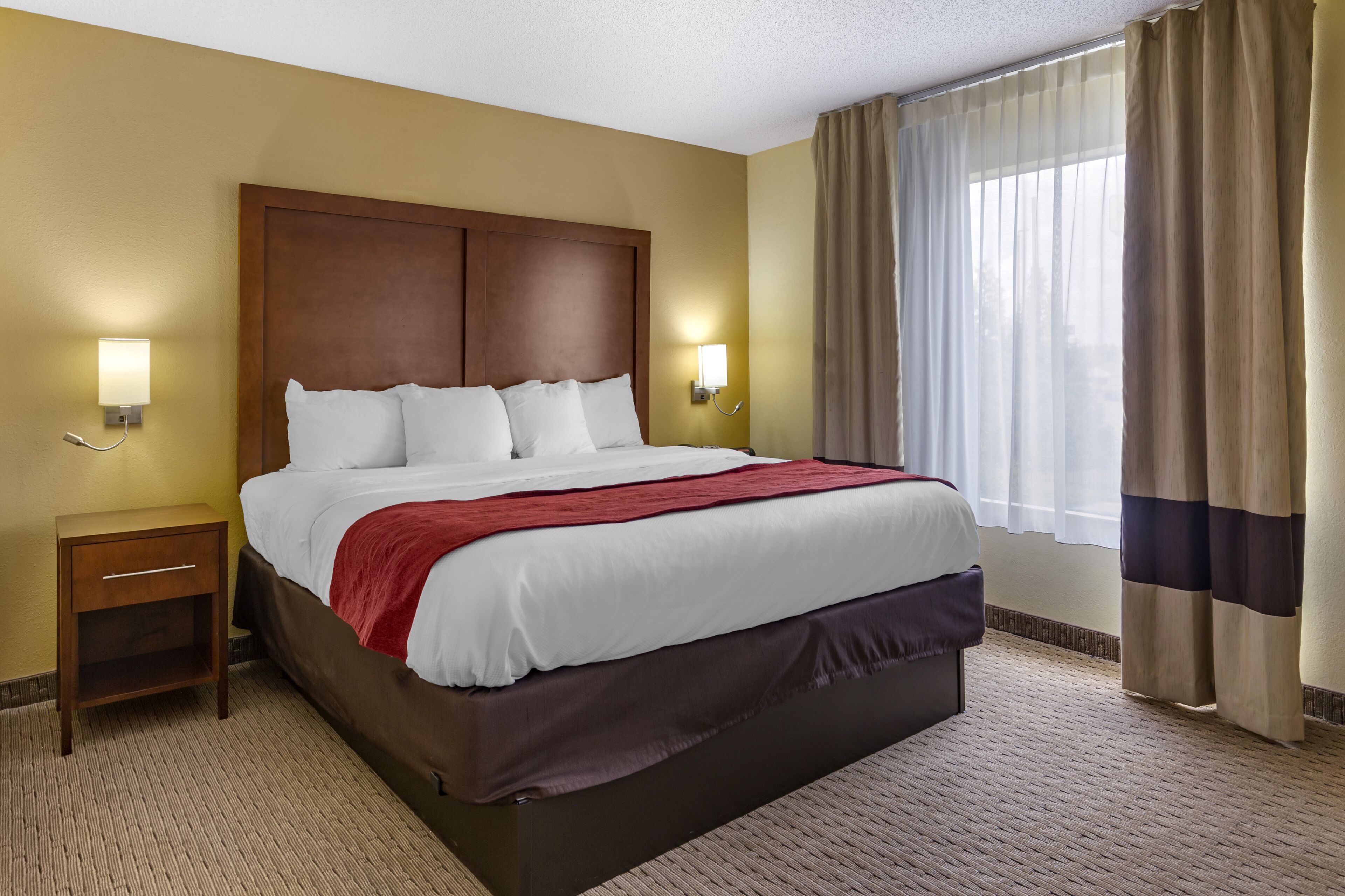 A hotel room with a large bed, two bedside tables, a wooden headboard, and a window with curtains.