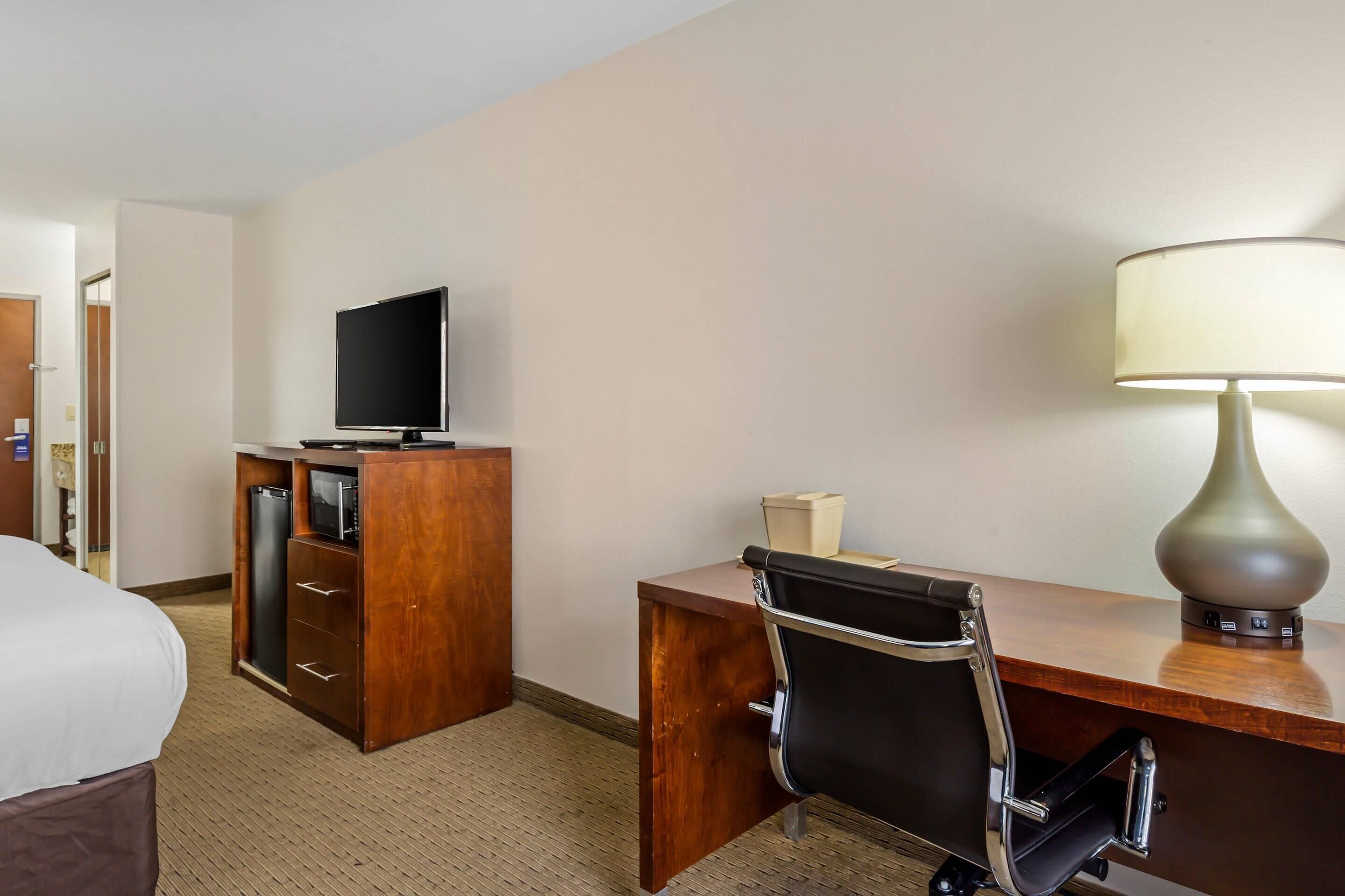 A hotel room with a bed, a desk with a chair, a television, a lamp, and a minibar.