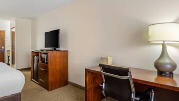 A hotel room with a bed, a desk with a chair, a television, a lamp, and a minibar.