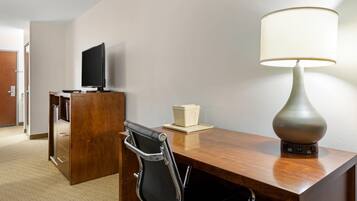 A hotel room with a wooden desk, a black office chair, a television on a stand, a lamp, and a small table with a box.