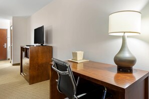 A hotel room with a wooden desk, a black office chair, a television on a stand, a lamp, and a small table with a box.