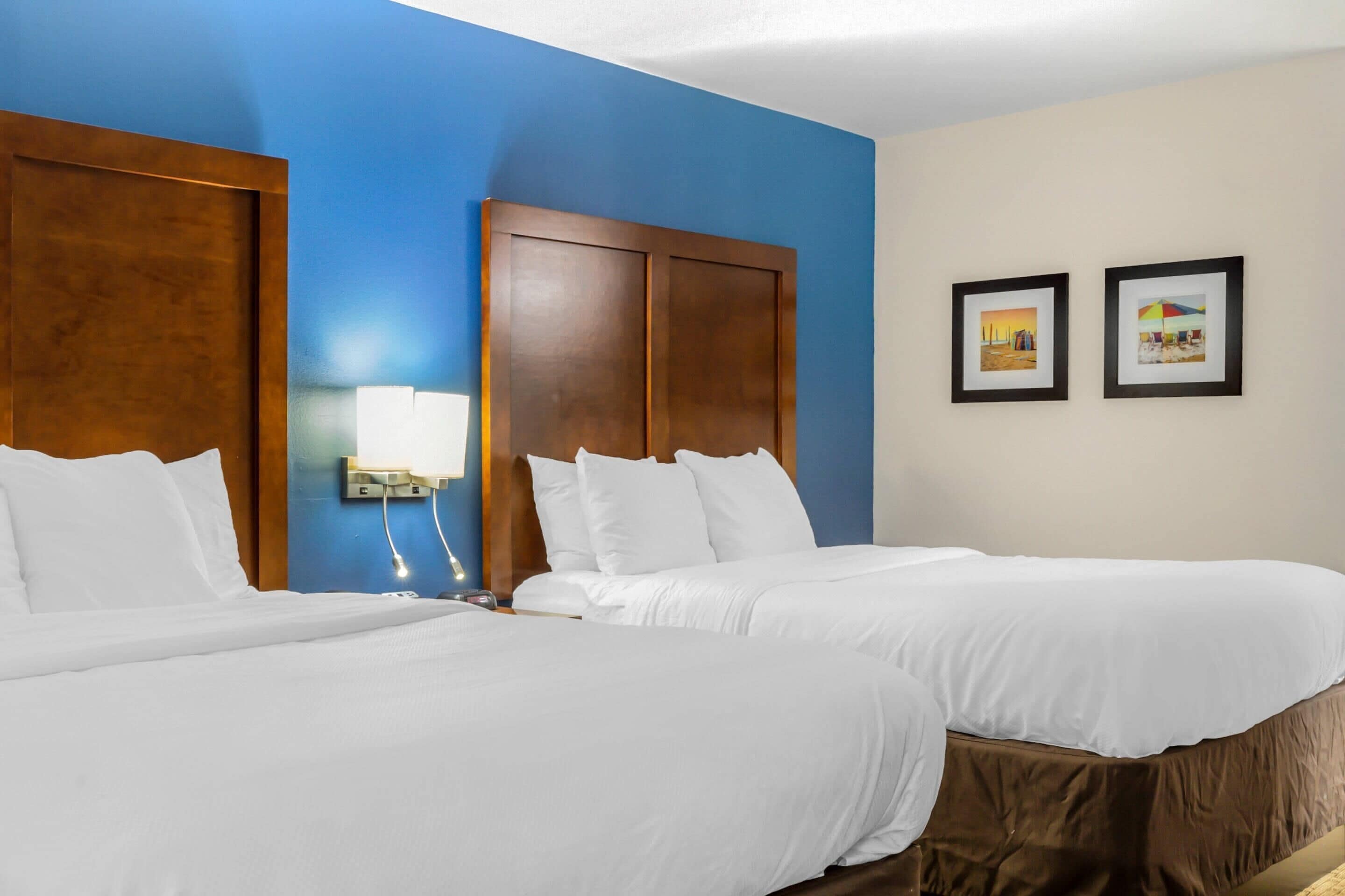 A hotel room with two beds, a wooden headboard, a nightstand, and a wall-mounted lamp.