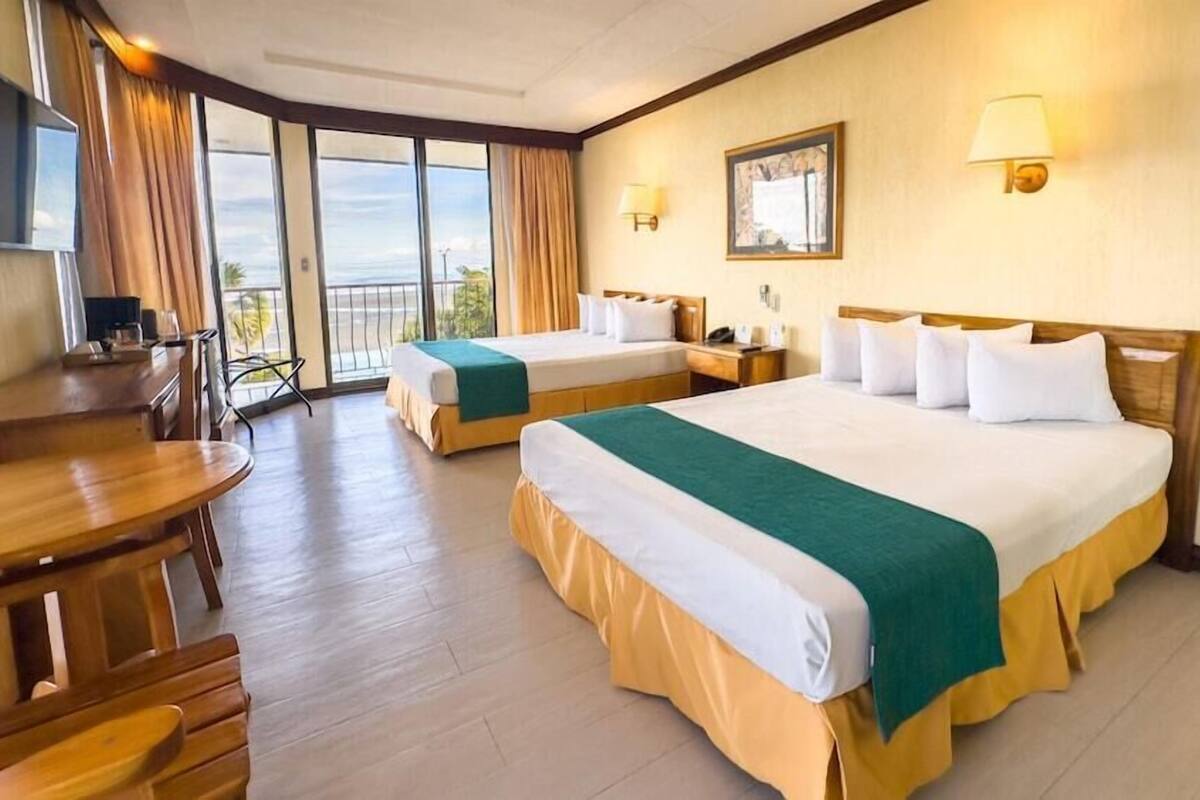 A hotel room with two beds, a desk, a chair, a TV, and a balcony with curtains.