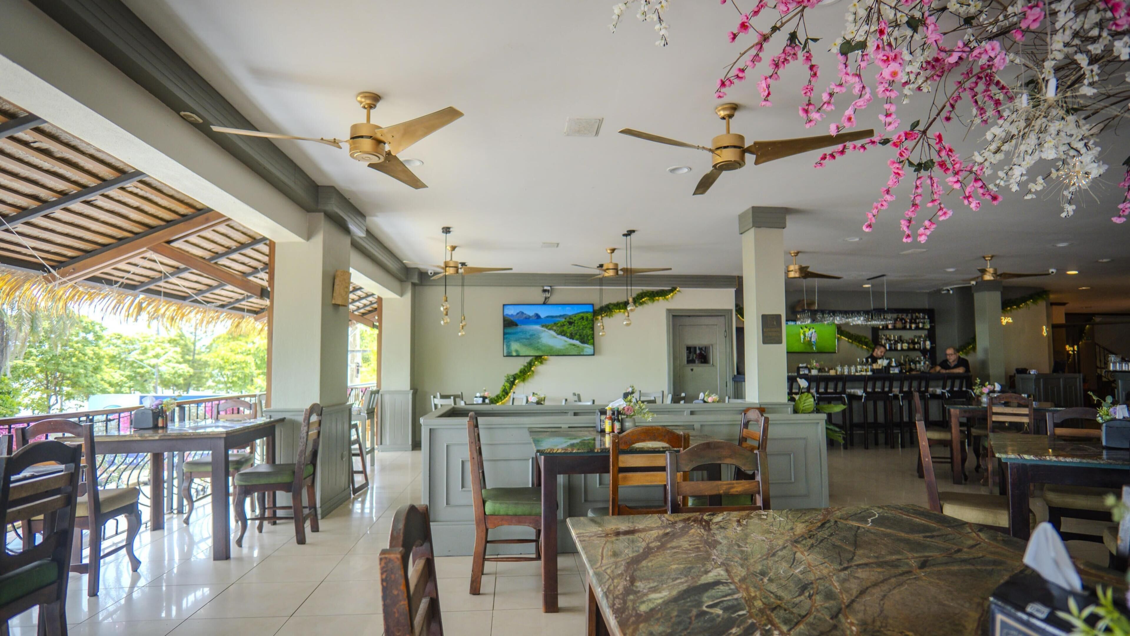 Un restaurante con zona de bar, varios ventiladores de techo y una pantalla de televisión mostrando un paisaje.